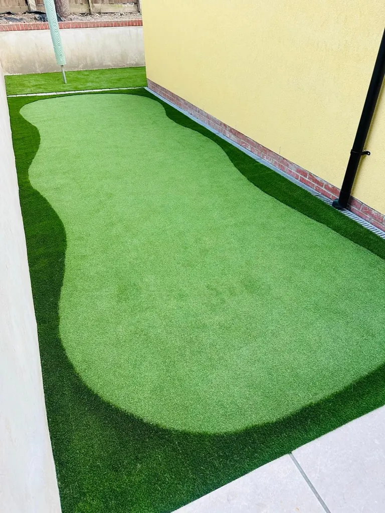 Saygrass putting-green grass in a curved shape, surrounded by darker landscape artificial grass beside a yellow house wall