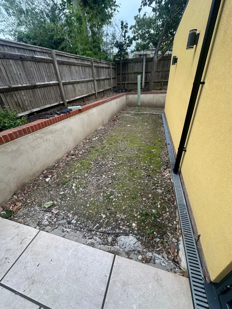 Narrow side garden before installation, with bare ground beside a yellow wall and drainage channel